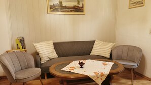 A living room with a brown sofa, two grey armchairs, a wooden coffee table with a floral tablecloth, and two framed pictures on the wall. - Peper, 2-room apartment, below, 2 P. - Peper, Ute (Neustadt in Holstein)