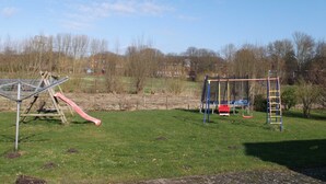 A playground with a slide and swings in a grassy area with buildings in the background. - Peper, 2-room apartment, below, 2 P. - Peper, Ute (Neustadt in Holstein)