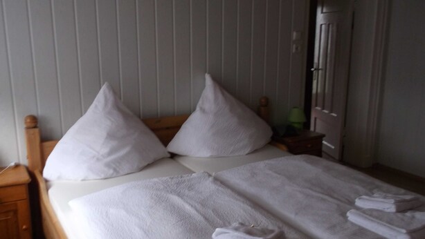 A neatly made bed with white linens and pillows, a wooden nightstand, and a door in the background. - Peper, 2-room apartment, below, 2 P. - Peper, Ute (Neustadt in Holstein)