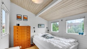 Ein Schlafzimmer mit einem Holzkoffer, einem Bett mit weißer Bettwäsche, zwei Fenstern mit Blick auf Grünflächen und einer Decke mit Holzbalken.