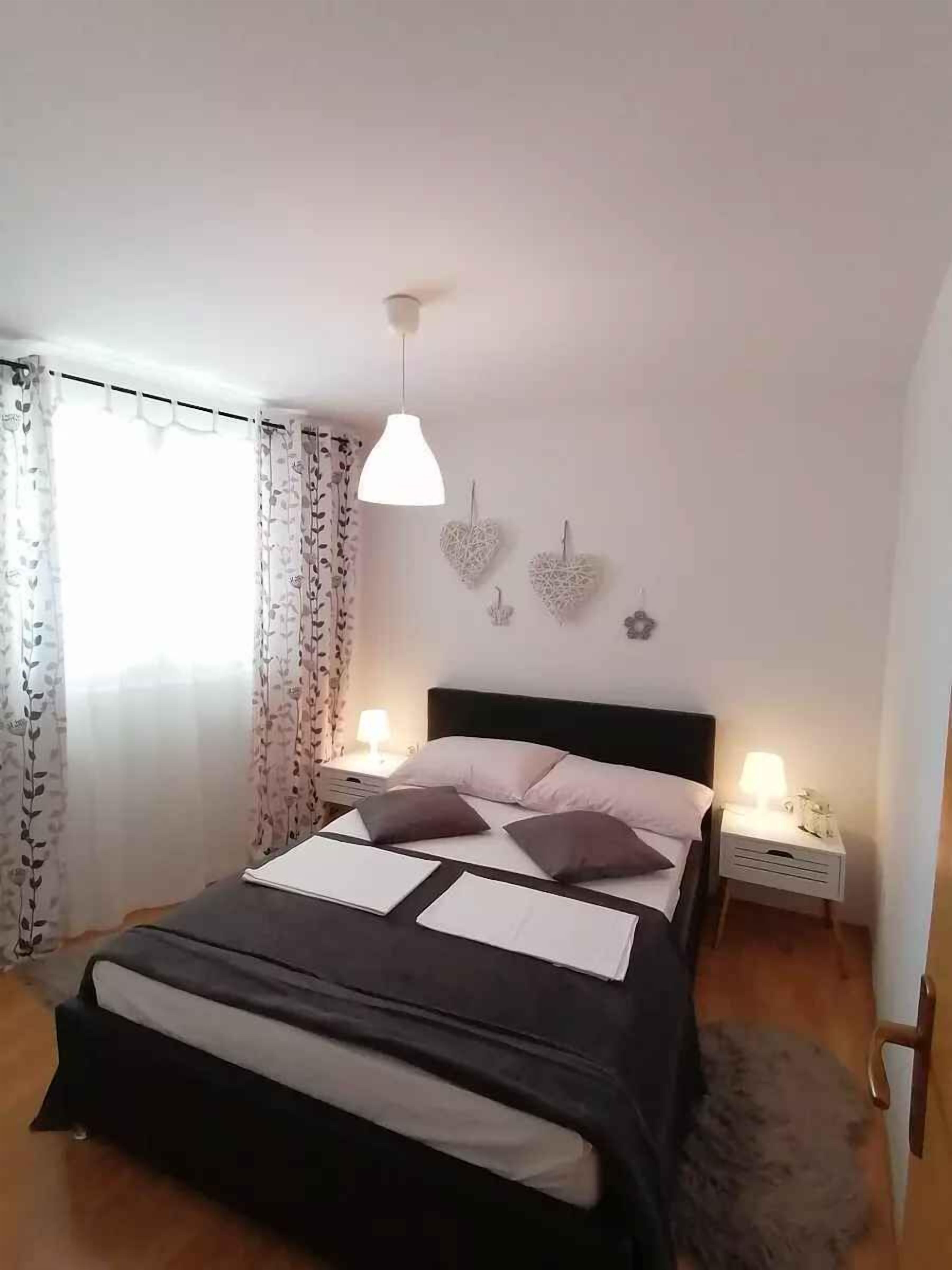 Una cama bien hecha con cabecera negra, dos lámparas de noche, decoraciones de pared con forma de corazón y una ventana con cortinas florales.