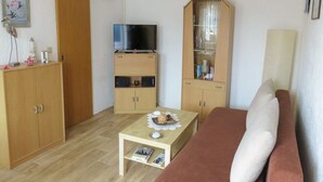 A compact living space with a sofa, a TV on a cabinet, a small table with a book, and a cabinet with glass doors. - Bungalow Alms, Germany (Ribnitz-Damgarten)