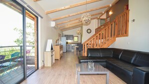 A spacious living area with a black leather sofa, a wooden staircase, and a kitchen in the background.