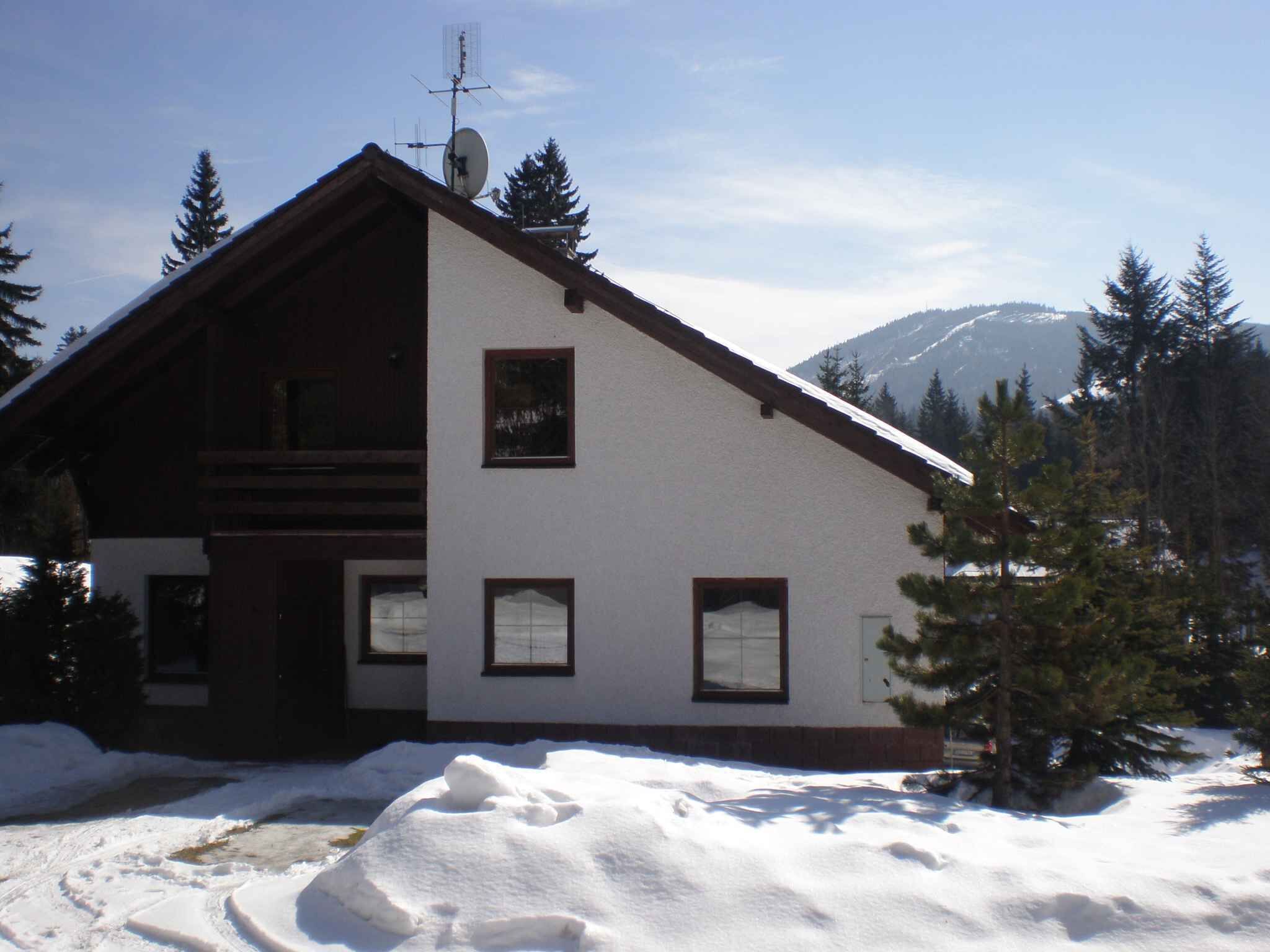 A two-story house with a sloped roof, surrounded by snow and trees, with a mountain in the background.