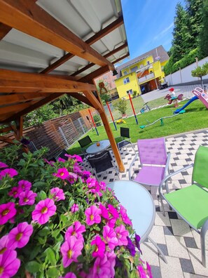 Een overdekt terras met een houten pergola, zitplaatsen buiten en kleurrijke bloemen.