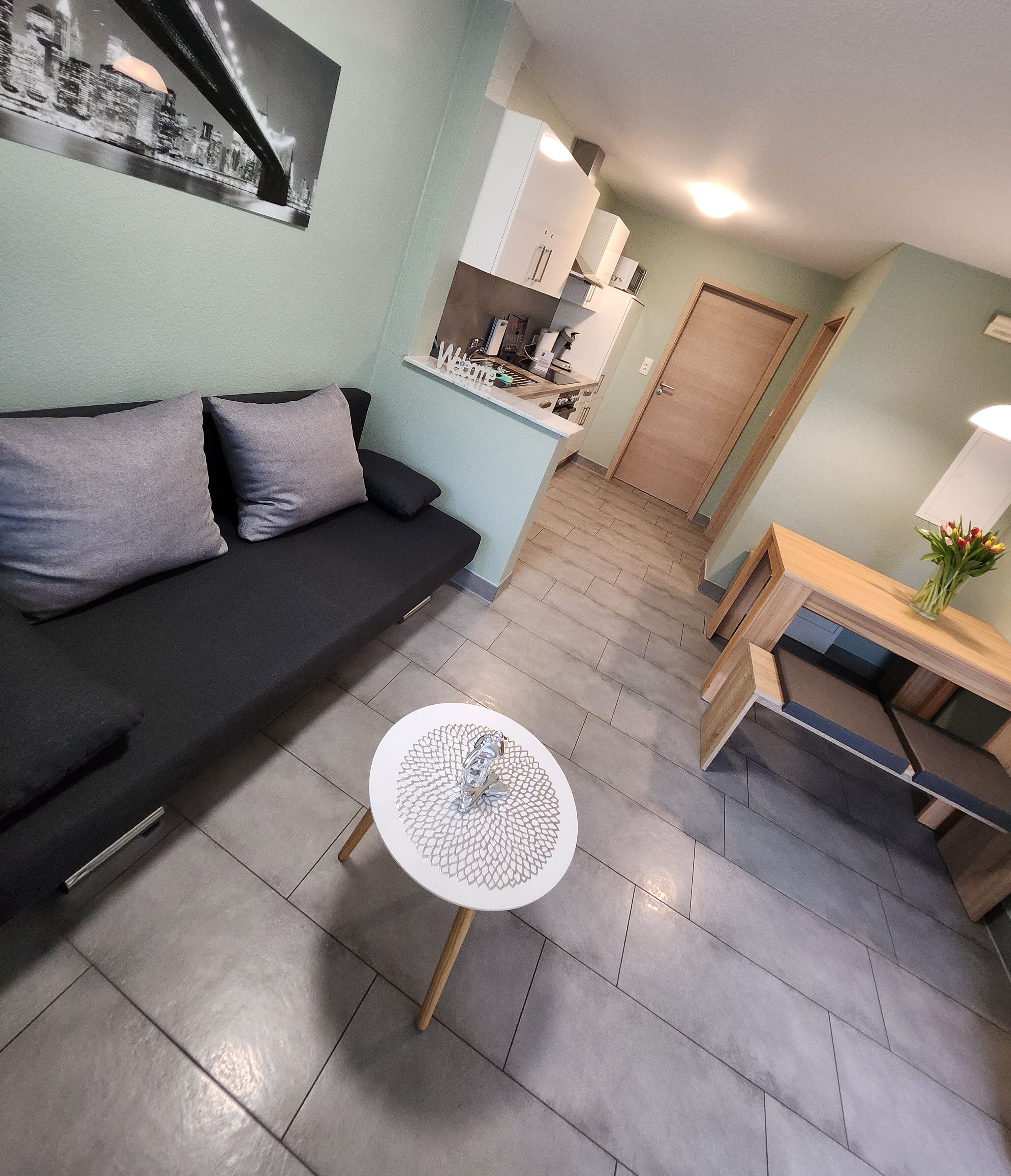 A modern living room with a black sofa, a round white table, and a kitchen area in the background.