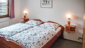 Una cama doble con edredón estampado, dos mesitas de noche con lámparas, un cuadro enmarcado en la pared y una ventana con cortinas.