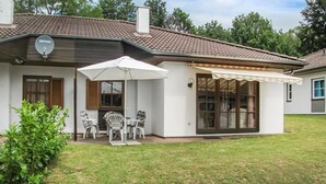 Una casa con patio amueblado con una mesa y sillas bajo un paraguas blanco, antena parabólica en el techo y un ventanal con cortinas.