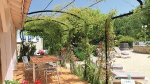 Une terrasse couverte, agrémentée d’une table et de chaises, entourée d’une végétation luxuriante et de plantes en fleurs.