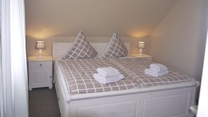 A neatly made bed with a checkered bedspread and two pillows, two bedside lamps, and a white nightstand with a drawer. - Cottage Riedensee (Kühlungsborn)