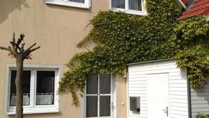 A two-story house with a red roof, white windows, and a door with a glass panel. There is a wooden bench and a tree in front of the house. - Cottage Riedensee (Kühlungsborn)