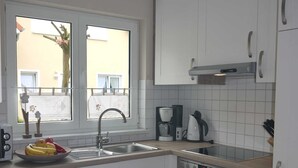 A modern kitchen with white cabinets, a built-in oven, a coffee maker, and a window with a view of the outside. - Cottage Riedensee (Kühlungsborn)