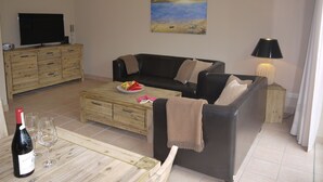 A living room with a black leather sofa, a wooden coffee table with a fruit bowl, a television on a wooden cabinet, and a painting on the wall. - Cottage Riedensee (Kühlungsborn)