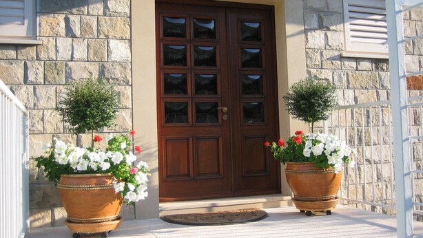 A stone building with a wooden double door, two potted plants with white and red flowers, and a white metal railing. - Apartment in Dubrovnik with swimming pool and sea view   (A2) (Dubrovnik Mokosica)