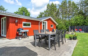 A red wooden building with a patio area featuring a table and chairs, a barbecue grill, and a small garden with potted plants. - Nice home in Oksbøl with WiFi (Oksbøl)
