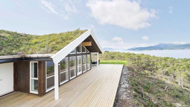 Ein modernes Haus mit großen Fenstern und einer Holzterrasse mit Blick auf einen See und Berge.