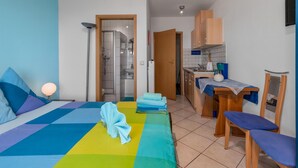 Een slaapkamer met een bed, nachtkastje en een klein tafeltje. Er is een badkamer met een douche en toilet. De kamer heeft een raam en een deur naar de keuken.