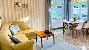 A living room with a sofa, a coffee table, a dining table with chairs, a lamp, and a window with curtains. - Apartment Schwalbennest - House Gundelrebe (Bergen auf Rügen)