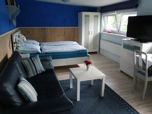 Una habitación con una cama, un sofá, una mesita con un florero, un escritorio con televisor y una ventana.