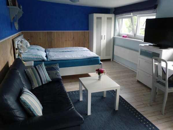 A room with a bed, a sofa, a small table with a vase, a desk with a TV, and a window. - Apartment Schwalbennest - House Gundelrebe (Bergen auf Rügen)