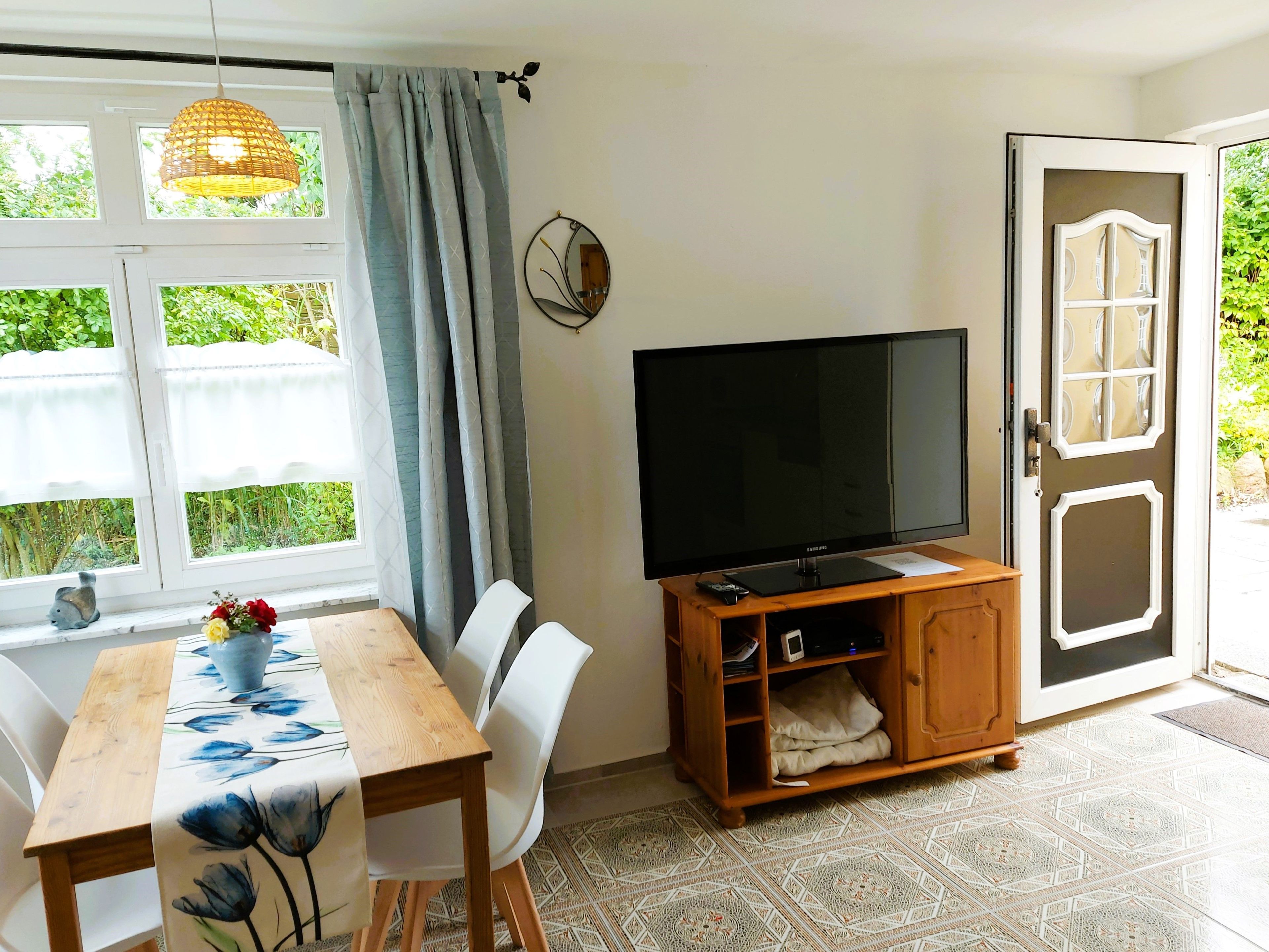 A room with a dining table, chairs, a television on a wooden stand, a window with curtains, and a door leading outside.
