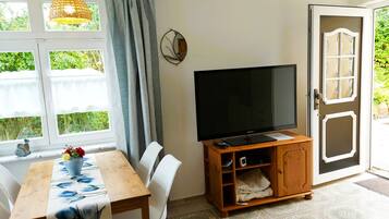 A room with a dining table, chairs, a television on a wooden stand, a window with curtains, and a door leading outside.