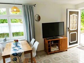 Een kamer met een eettafel, stoelen, een televisie op een houten dressoir, een raam met gordijnen en een deur naar buiten.