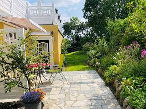 Un sendero conduce a un edificio de fachada amarilla, una reja blanca y un jardín con diversas plantas y flores.