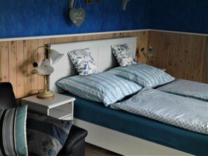 A bedroom with a wooden headboard, a bedside table with a lamp, two pillows, and a bed with a blue bedspread.