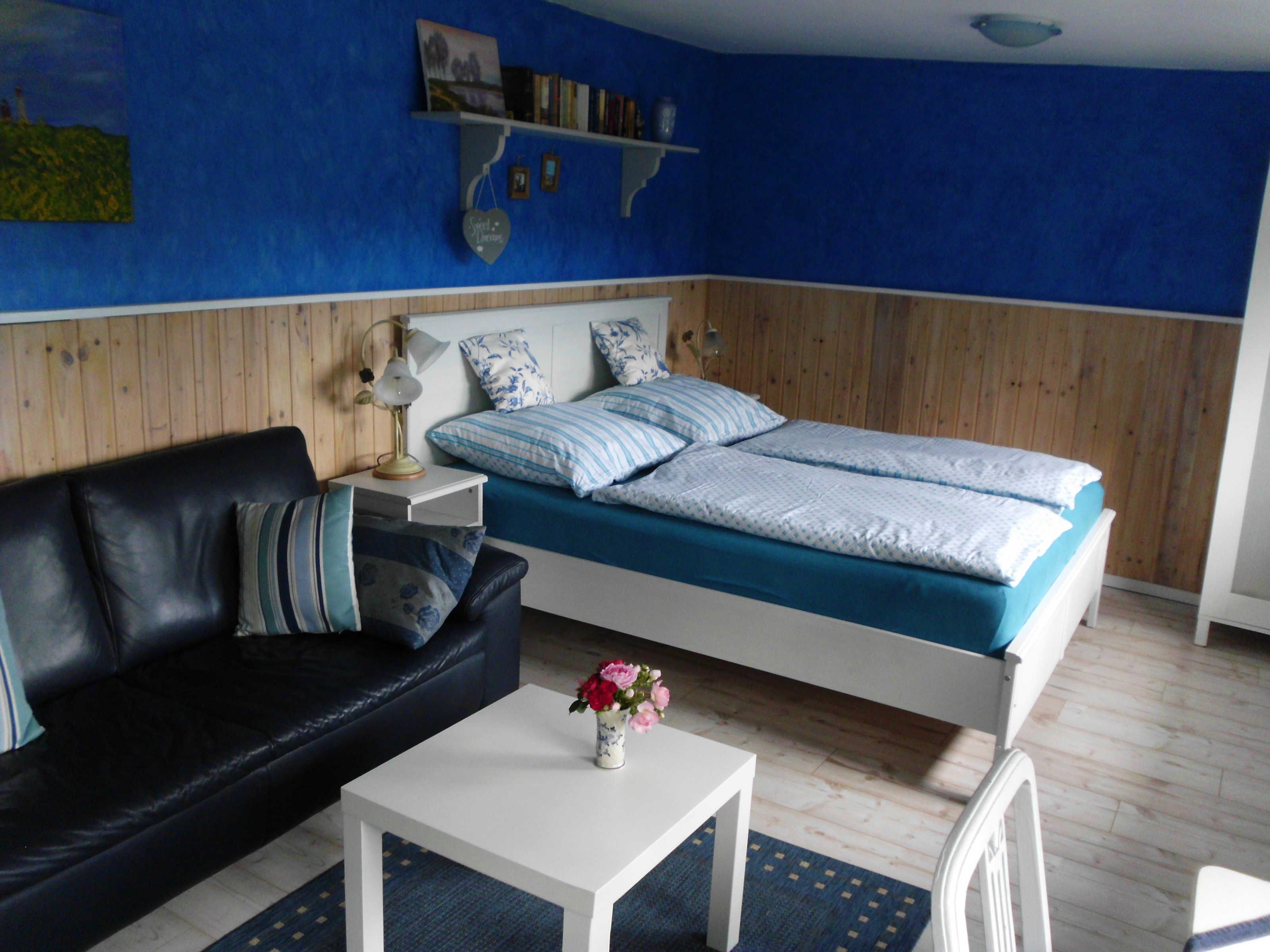 A bedroom with a bed, a black leather sofa, a white table, and a small vase with flowers.