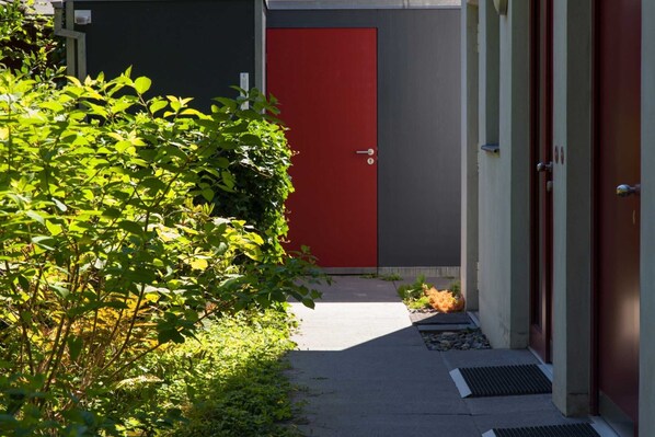 A narrow pathway leading to a red door, flanked by greenery and a concrete wall.