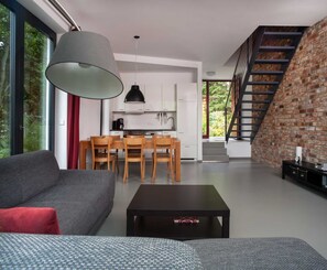A modern living room with a grey sofa, a black coffee table, and a dining area with wooden chairs and a table. There is a kitchen with white cabinets and a black pendant light. A brick wall and a staircase are visible in the background.