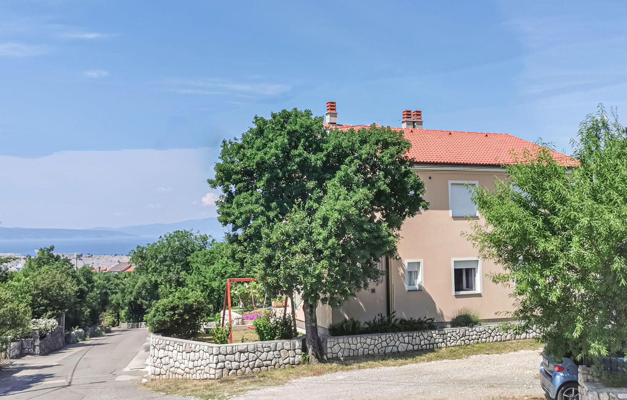 Una casa a due piani con tetto in tegole rosse, immersa nel verde e circondata da un muro in pietra.
