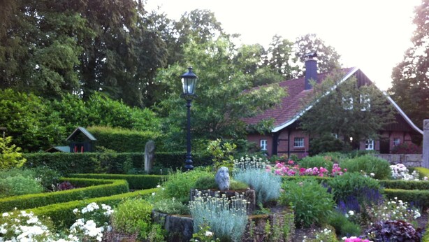 Ein Garten mit verschiedenen Blumen, einer Steinskulptur und einem traditionellen Haus im Hintergrund.