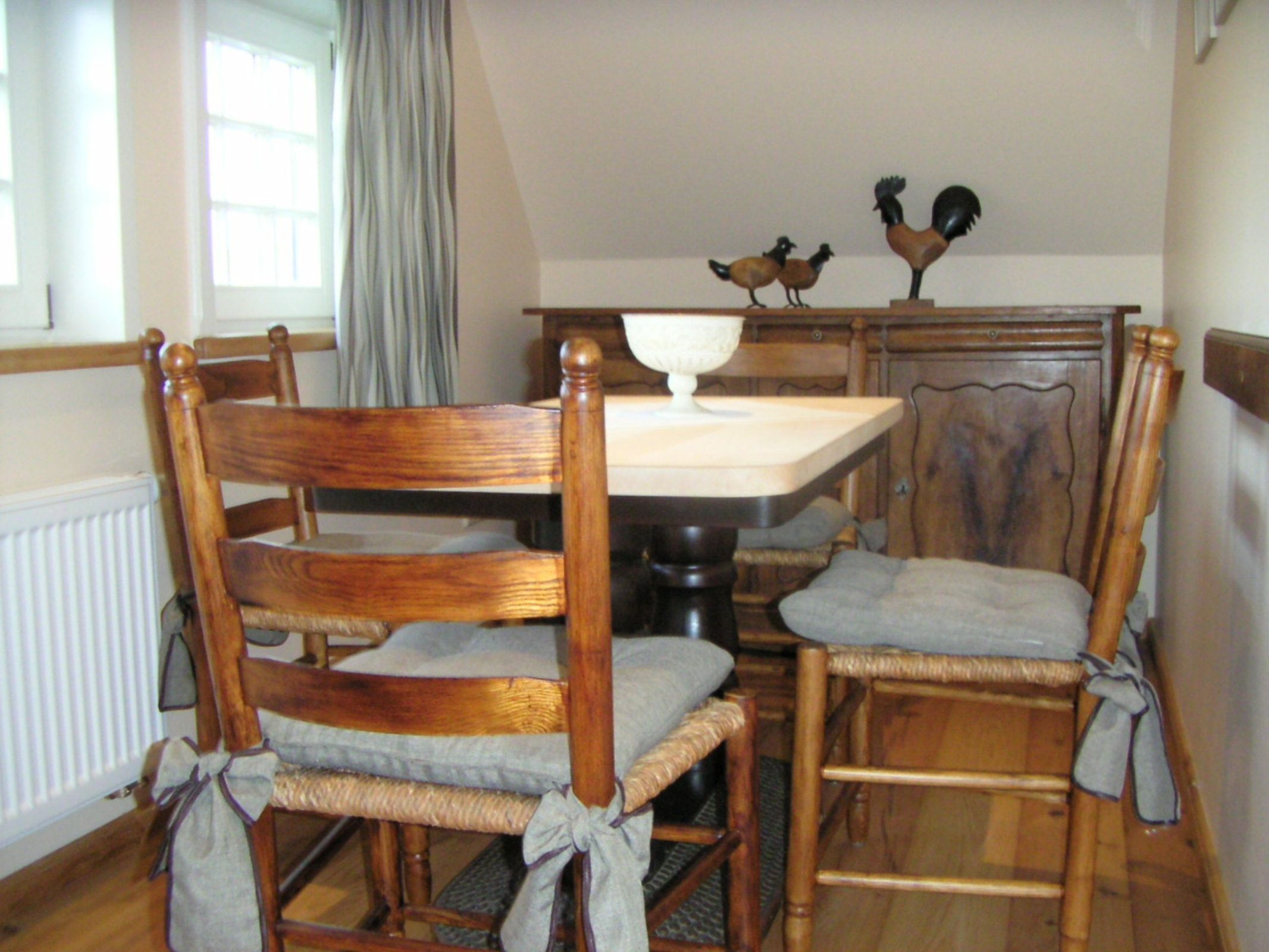 Un coin repas comprenant une table et des chaises, un radiateur, une fenêtre avec des rideaux et un meuble en bois agrémenté d’objets décoratifs.