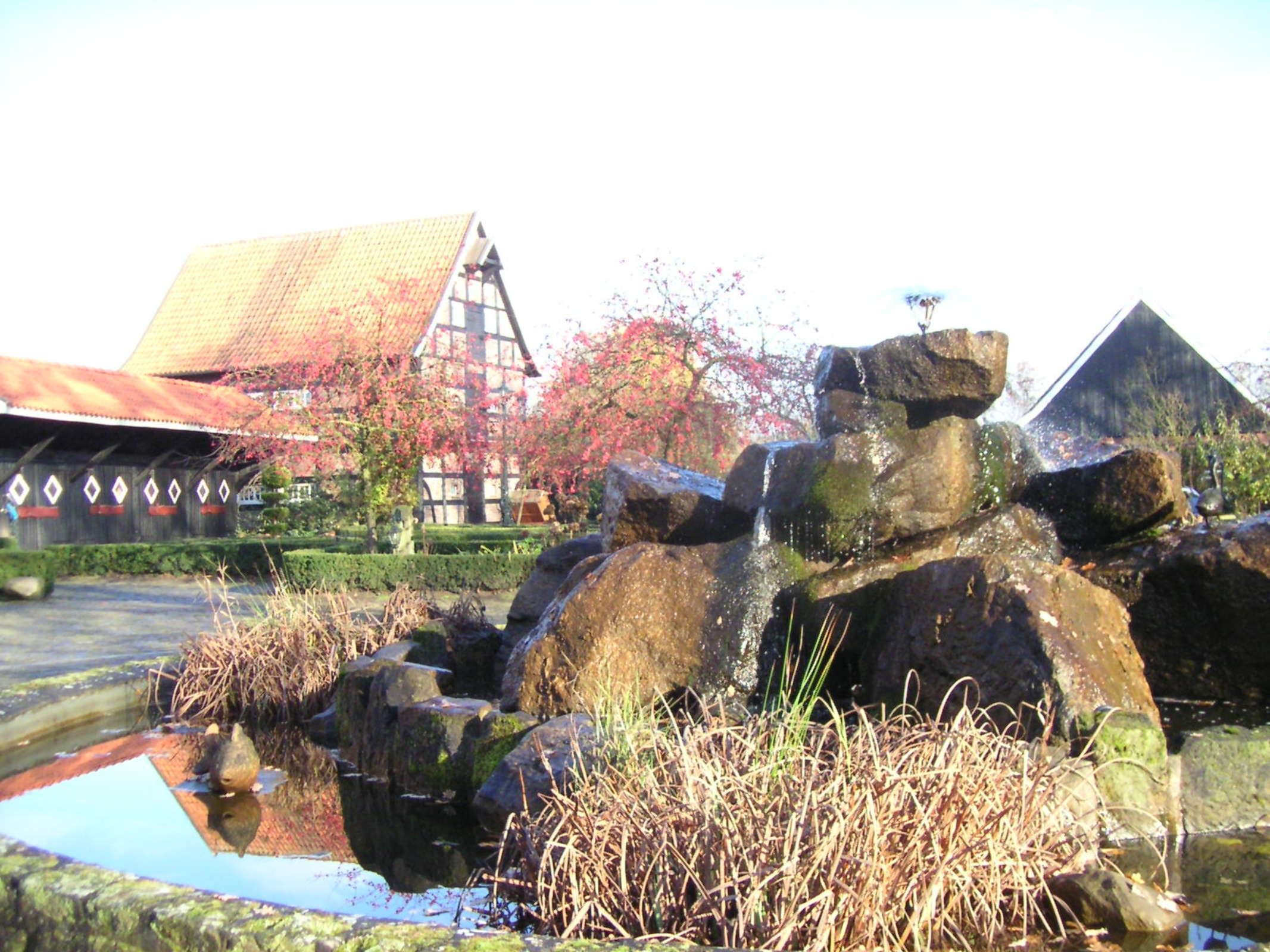 A traditional building with a sloped roof, a stone structure with water flowing over it, and a garden with autumn foliage.