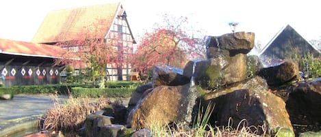 A traditional building with a sloped roof, a stone structure with water flowing over it, and a garden with autumn foliage.