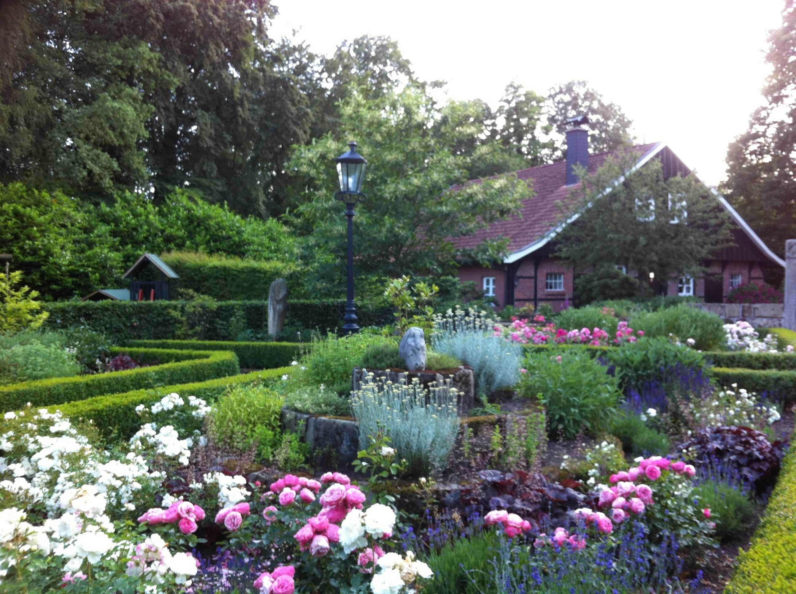 Un jardín con diversas flores, una escultura de piedra y una casa tradicional al fondo.