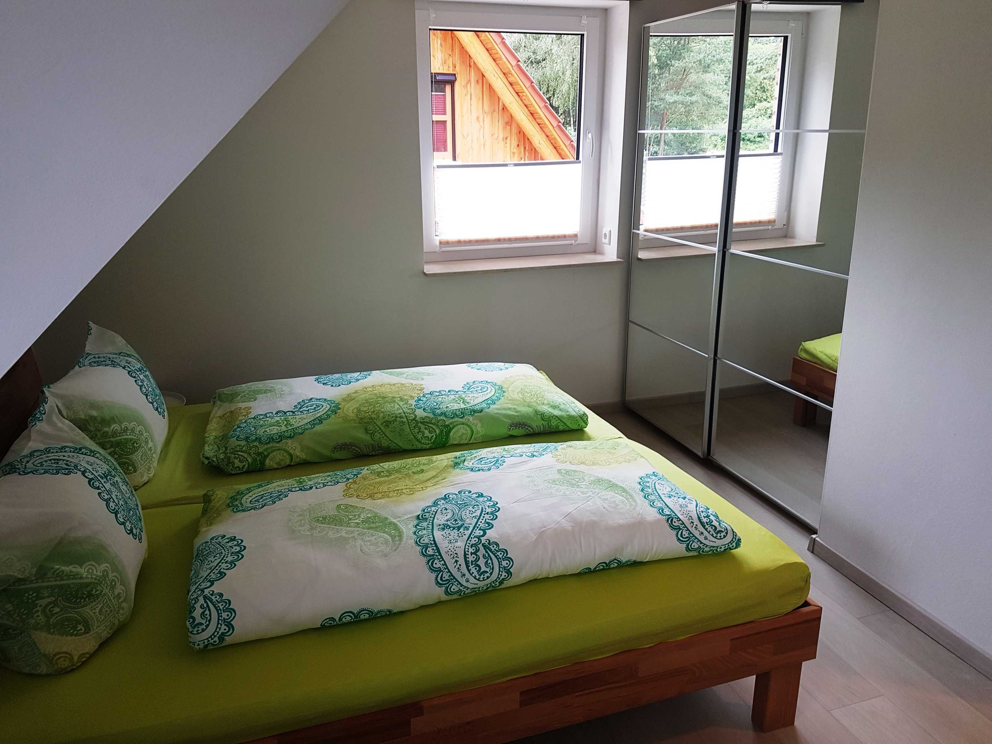 A bedroom with a bed, green and white patterned bedding, a wooden bench, and a window with a view of a wooden structure outside.