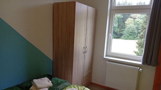 A bedroom with a bed, a wardrobe, a window with a view of greenery, and a radiator.