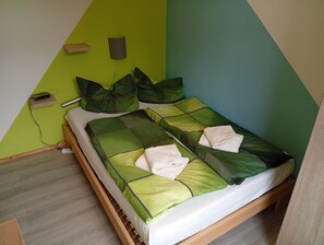 A single bed with green bedding and pillows, a bedside table with a lamp, and a wall-mounted power outlet. - 3-Person Apartments - Apartment 2 (Elend)