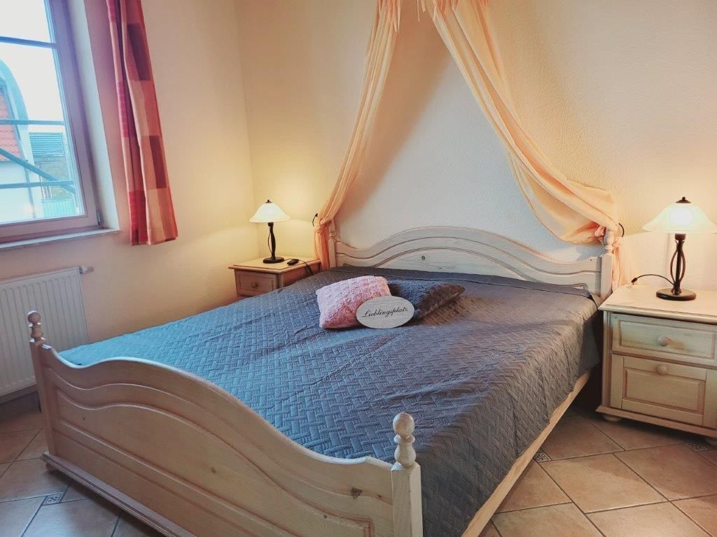 Ein Schlafzimmer mit einem Bett, zwei Nachttischen, einem Fenster mit Vorhängen und einem Beistelltischchen mit einer Lampe.