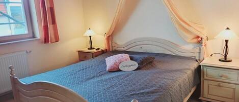 Ein Schlafzimmer mit einem Bett, zwei Nachttischen, einem Fenster mit Vorhängen und einem Beistelltischchen mit einer Lampe.
