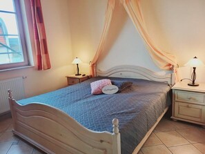 Quarto com cama, duas mesinhas de cabeceira, uma janela com cortinas e um criado-mudo com abajur.