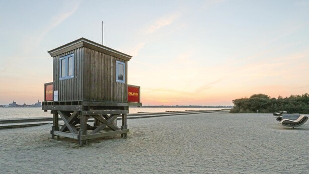 Ein hölzerner Rettungsschwimmturm mit Fahnenmast und zwei roten Schildern an der Seite, gelegen an einem sandigen Strand mit Blick auf eine entfernte Stadtlandschaft bei Sonnenuntergang.