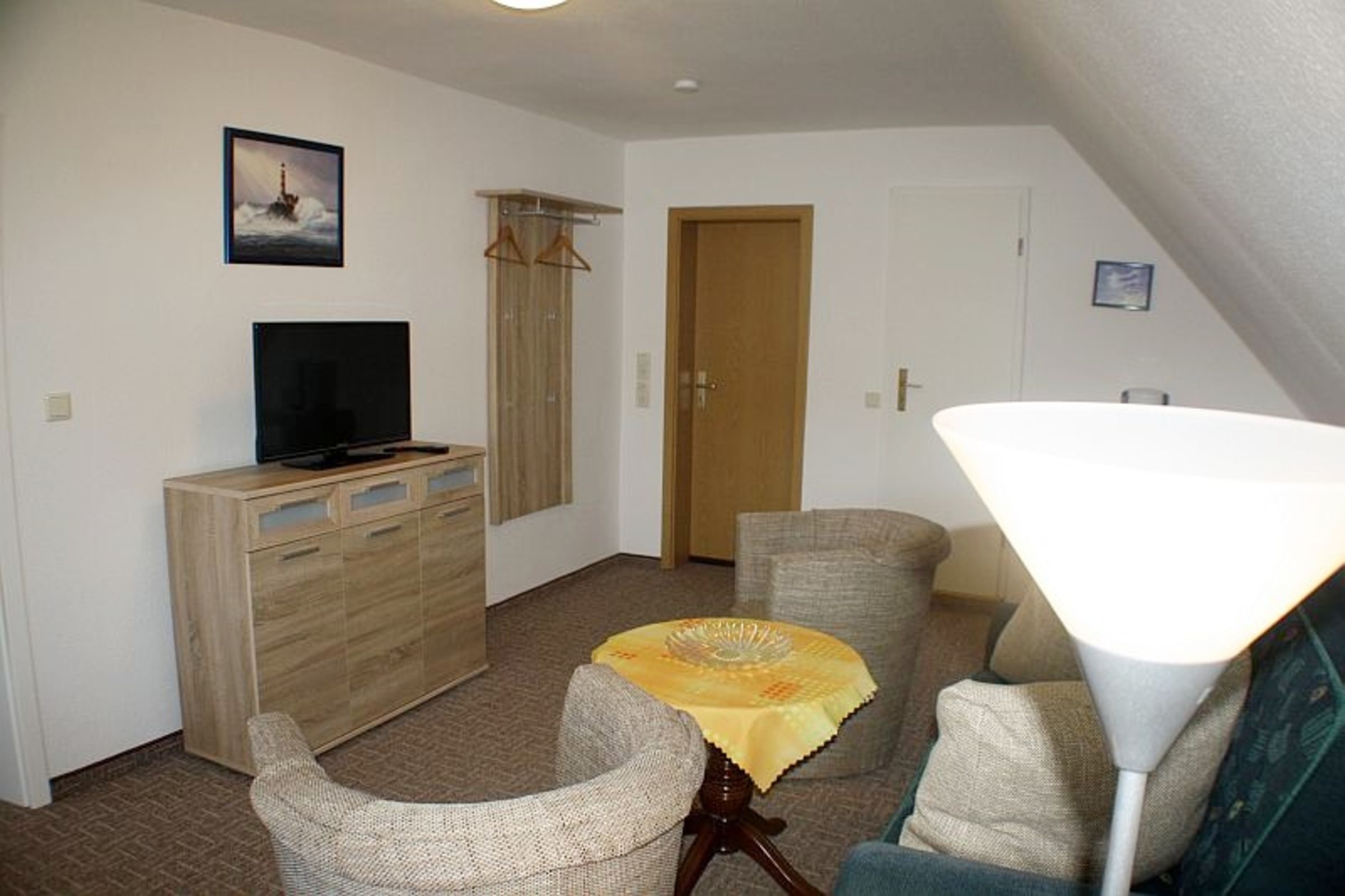 Una sala de estar con televisión, un mueble de madera, una mesa con mantel amarillo, dos sillones, una lámpara y una puerta.