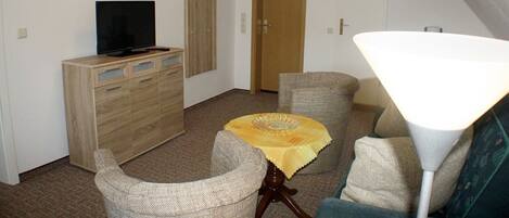Una sala de estar con televisión, un mueble de madera, una mesa con mantel amarillo, dos sillones, una lámpara y una puerta.