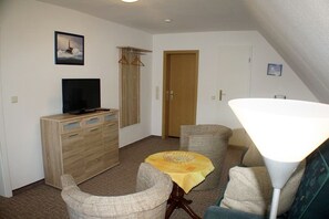 Un salon comprenant une télévision, un meuble en bois, une table recouverte d’une nappe jaune, deux fauteuils, une lampe et une porte.