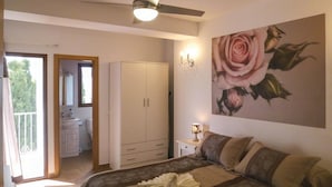 A bedroom with a bed, a wardrobe, a painting of a rose, a ceiling fan, and a bathroom visible through an open door.