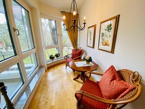 A room with a wooden floor, a dining table with chairs, a wicker sofa, a chandelier, and large windows with curtains.